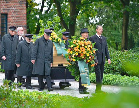 Szene einer Bestattung, bei der eine Gruppe Menschen den mit Blumen geschmückten Sarg über den Friedhof geleiten. Das Bild verlinkt auf die Seite über Erdbestattungen.
