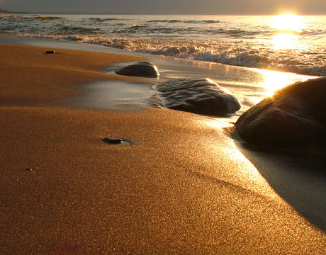 Ausschnitt eines Strands bei Sonnenuntergang. Das Meer berührt sanfte geformte Steine am Strand. Das Bild verlinkt auf die Seite über Seebestattungen.