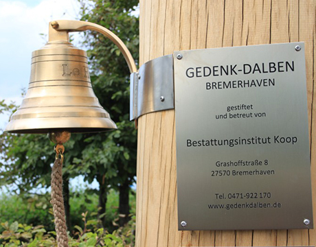 Foto von einem Gedenk-Dalben. Man sieht eine Glocke und eine Plakette mit der Aufschrift: Gedenk-Dalben Bremerhaven, gestiftet und betreut von Bestattungsinstitut Koop.