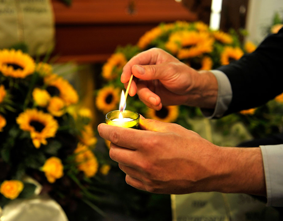 Zwei Hände ragen ins Bild. Eine hält ein Teelicht, die andere ein brennendes Streichholz und zündet das Teelicht an. Im Hintergrund sind Gestecke aus Sonnenblumen zu erkennen.